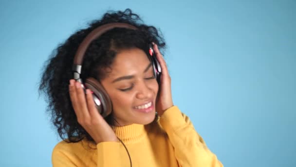 Modelo de moda sorridente 4K com fones de ouvido posando e dançando contra fundo azul em estúdio. Bonito jovem mestiço de raça africana modelo feminino apreciando música — Vídeo de Stock