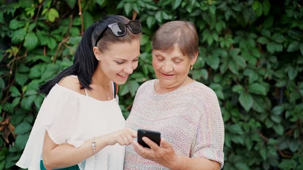 Granddaughter shows old grandmother something in smartphone, teaches her to handle with modern gadget and technology. Family, love, care concept. — Stock Photo, Image