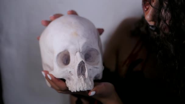 Страшный портрет молодой девушки с макияжем крови Хэллоуина, держащей настоящий череп. Красивая латинская женщина с вьющимися волосами позирует в студии. Медленное движение — стоковое видео
