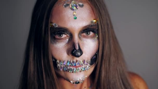 Молодая девушка с творческим Хэллоуин лицо искусства в студии с серым фоном. Портрет гламурного черепа со стразами и блестками. Профессиональный макияж для праздника. Медленное движение . — стоковое видео