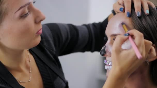 Maskenbildner bemalt Fettfarbe für Halloween im Atelier. Frau zeichnet einen glamourösen Totenkopf mit Strass und Pailletten auf ein schönes junges Mädchen mit langen Haaren. Zeitlupe. — Stockvideo