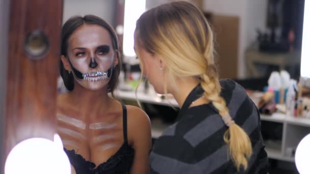 Wizażystka farby szminki na Halloween w studio. Woman rysowanie efektowne czaszki z dżetów i cekiny na piękna młoda dziewczyna z długimi włosami. Odbicie w ruchu Mirror.Slow — Wideo stockowe