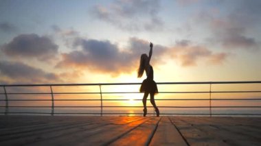 Balerin Bale tutu ve nokta güneş doğarken setin okyanus ya da deniz plaj yukarıda üzerinde dans ediyor. Güzel sarışın genç kadın uzun saçlı uzanan pratik ve egzersizleri silüeti. Ağır çekim.