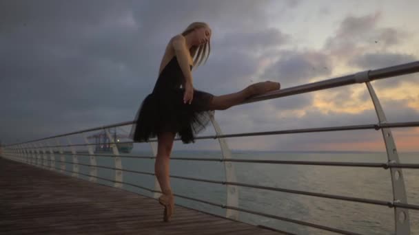 Tanzende Ballerina in schwarzem Balletttutu und Spitze auf einem Damm über dem Meer bei Sonnenaufgang oder Sonnenuntergang. junge attraktive blonde Frau mit langen Haaren übt Stretching und Übungen. Zeitlupe. — Stockvideo