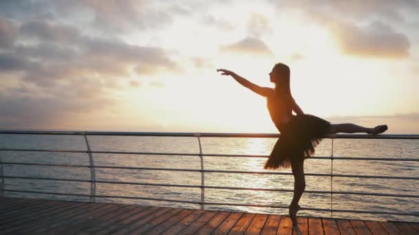 Танцююча балерина в чорному балеті навчає і вказує на набережну над океаном або морем на заході сонця. Силует молодої красивої блондинки з довгим волоссям практикує розтягування і вправи. СЛЮТ Мочіон . — стокове відео