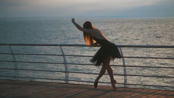 Bela cena de bailarina dançante em tutu de balé preto e ponto em aterro acima do mar ao nascer do sol ou pôr do sol.Mulher loira bonita com cabelos longos praticando alongamento e exercícios. LOW MOTION . — Vídeo de Stock