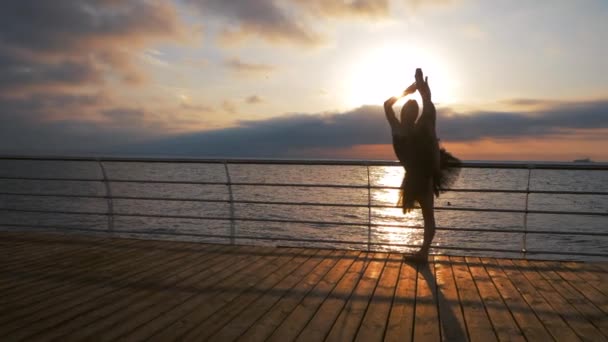 Schöne Szene einer tanzenden Ballerina in schwarzem Balletttutu, die bei Sonnenaufgang auf eine Böschung über Meer oder Strand zeigt. Silhouette der Frau hebt Bein über Kopf. Zeitlupe. — Stockvideo