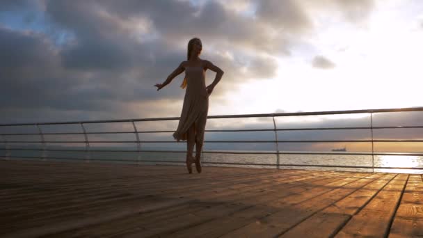 Танцююча балерина в бежевому шовковому одязі і путе на набережній над океаном або морем на сході або заході сонця. Силует молодої жінки з довгим волоссям, що практикує класичні вправи з емоціями. . — стокове відео
