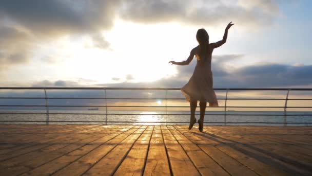 Bailarina bailarina en vestido de seda beige y punta en terraplén sobre el océano o el mar al amanecer o al atardecer. Silueta de mujer joven con pelo largo practicando ejercicios clásicos con emociones.MOVIMIENTO Lento . — Vídeo de stock