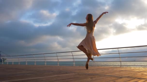 Танцующая балерина в бежевом шелковом платье и пуанты на набережной над океаном на восходе солнца. Молодая красивая блондинка с длинными волосами практикует растяжку и классические упражнения с эмоциями. . — стоковое видео