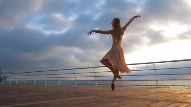 Dancing ballerina in beige silk dress and pointe on embankment above ocean at sunrise.Young beautiful blonde woman with long hair practicing stretching and classic exercises with emotions.SLOW MOTION. — Stock Video