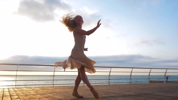 Bailarina bailarina en vestido de seda beige y punta en terraplén sobre el océano al amanecer. Joven mujer rubia hermosa con pelo largo practicando estiramientos y ejercicios clásicos con emociones. . — Vídeo de stock