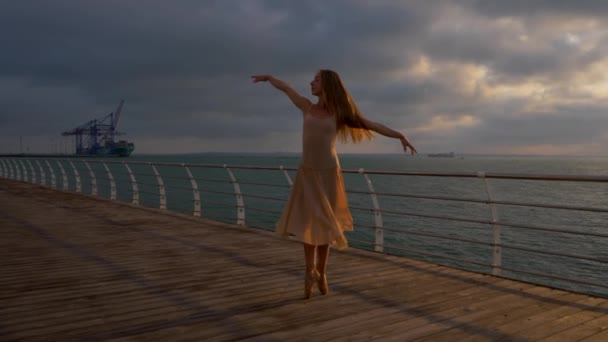 Tanzende Ballerina in beigem Seidenkleid und Spitze auf einem Damm über dem Meer oder am Strand bei Sonnenaufgang. junge schöne blonde Frau mit langen Haaren praktiziert klassische Übungen mit Emotionen. — Stockvideo