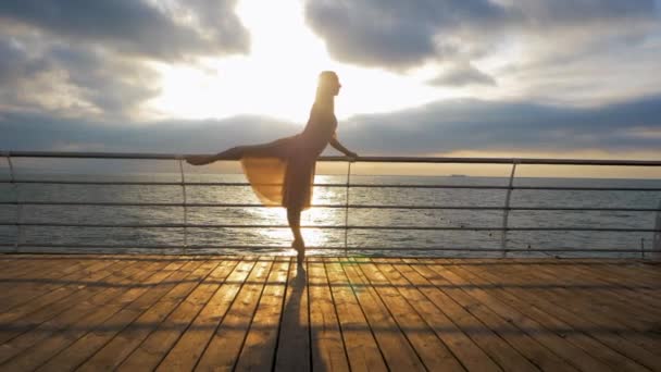 Танцююча балерина в бежевій шовковій сукні і путе на набережній над морським пляжем на сході або заході сонця. Силует молодої красивої блондинки з довгим волоссям практикує розтягування. СЛЮТ Мочіон . — стокове відео