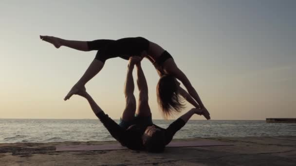 Silhouette di giovani coppie in forma che fanno acro-yoga sulla spiaggia del mare. Uomo sdraiato su piastre di cemento e bilanciamento donna nei suoi piedi. Bella coppia praticare yoga insieme . — Video Stock