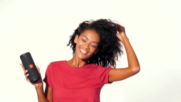 Joven hermosa mujer afroamericana disfrutando y bailando en el fondo blanco. Chica moderna de moda con peinado afro escuchando música por altavoz portátil inalámbrico . — Vídeos de Stock