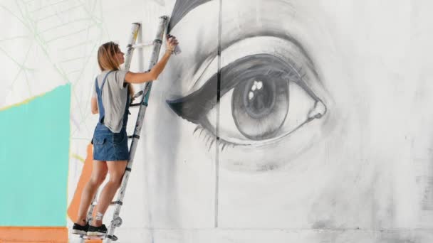 Krásná dívka, tvorba graffiti velké ženské tváře s Aerosolový sprej na městské ulici zdi. Ona stojí na žebříku. Výtvarného umění. Talentovaný student v džínové overaly kreslení obrázek