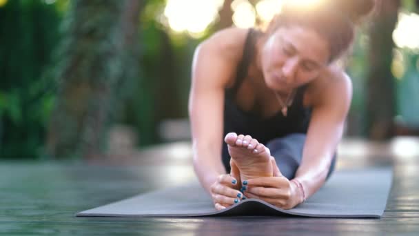 Estirar de cerca el pie. Chica haciendo torzal partido. Hanumanasana, pose de mono. Mujer de aspecto oriental joven practicando yoga en cubierta de madera en isla tropical . — Vídeos de Stock