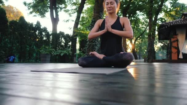 Сконцентрированная девушка сидит в позе лотоса с руками в намасте и медитирует или молится. Молодая женщина с восточным внешним видом практикует йогу в одиночку на деревянной палубе тропического острова — стоковое видео