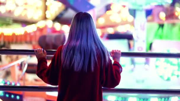 Dívka s odbarvenými vlasy modré stojící zpět do kamery a sledování rotace atrakcí v zábavním parku. Tisícileté bokovky životní styl. V noci, osvětlené město — Stock video