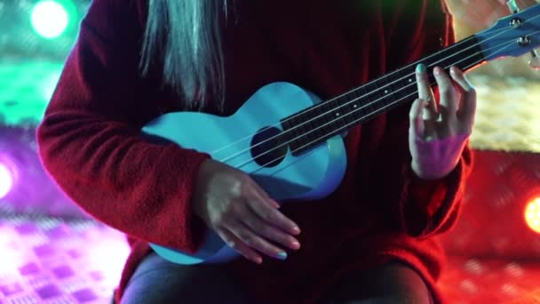 Mãos de menina bonita jovem jogando no ukulele azul enquanto sentado em escadas de néon brilhante no parque de diversões à noite — Vídeo de Stock