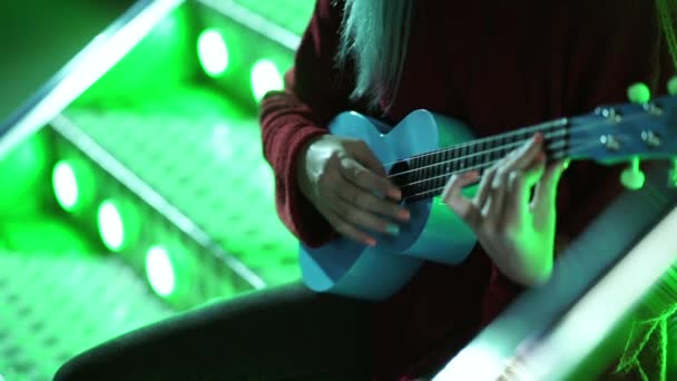 年轻漂亮的女孩的手玩蓝色四弦琴, 而坐在闪闪发光的霓虹灯楼梯在游乐园夜间 — 图库视频影像