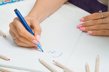 Oluşturun. El güzel mavi mektuplardan renkli kalemler çerçevesindeki tuval üzerine yazar. Hat komut dosyası. Mektup yazma sanatı. Eski beyaz tahta masada oturan bir bayan. Arka plan.