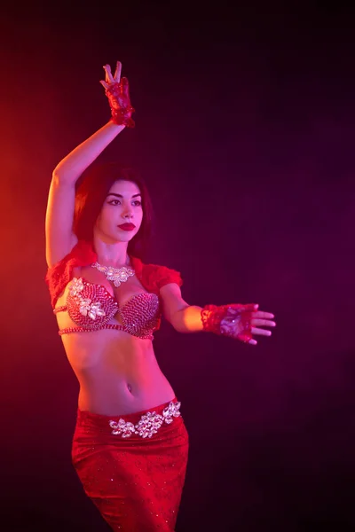 Woman in exotic shining red costume sexually moves her body. Sexy traditional oriental belly dancer girl dancing on neon smoke background. Muslims, temptation concept