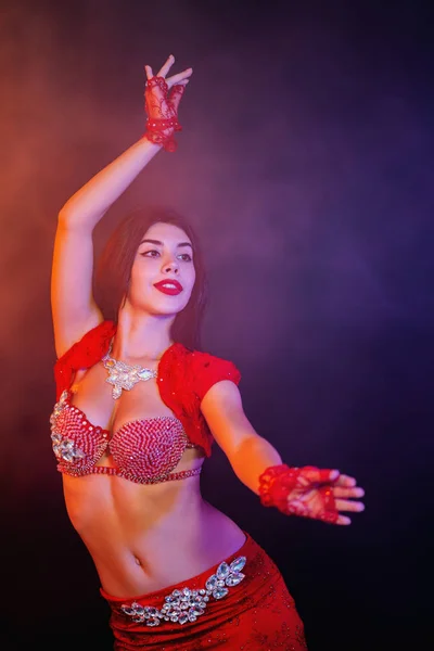 Tempting sexy traditional oriental belly dancer girl dancing on purple neon smoke background. Woman in exotic red costume sexually moves her semi-nude body.