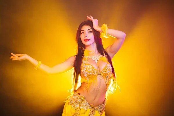 Séduisante danseuse orientale traditionnelle sexy du ventre dansant sur fond de fumée jaune néon. Femme en costume exotique avec des plumes bouge sexuellement son corps semi-nu . — Photo