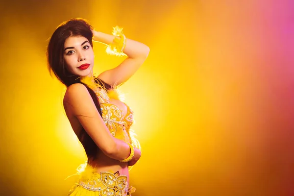 Alluring sexy traditional oriental belly dancer girl dancing on yellow neon smoke background. Woman in exotic costume with feathers sexually moves her semi-nude body — Stock Photo, Image