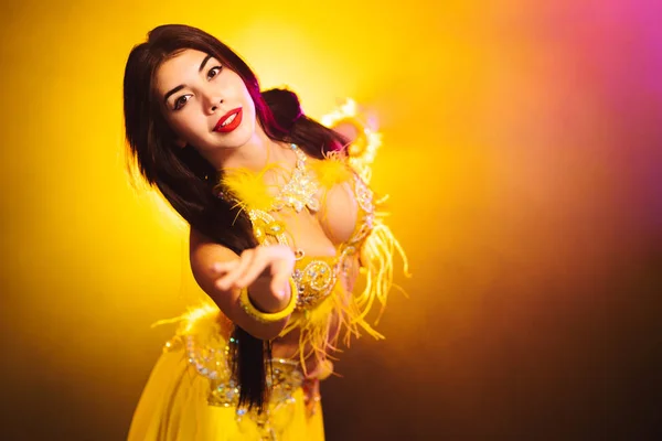 Tempting sexy traditional oriental belly dancer girl dancing on yellow neon smoke background. Woman in exotic costume with feathers sexually moves her semi-nude body — Stock Photo, Image