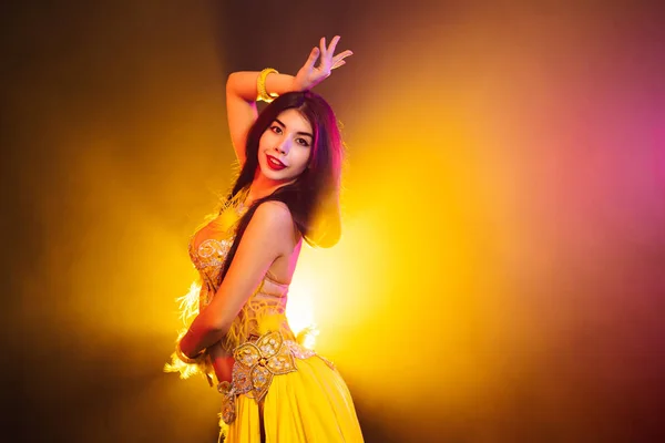 Seductive sexy traditional oriental belly dancer girl dancing on yellow neon smoke background. Woman in exotic costume with feathers sexually moves her semi-nude body. — Stock Photo, Image