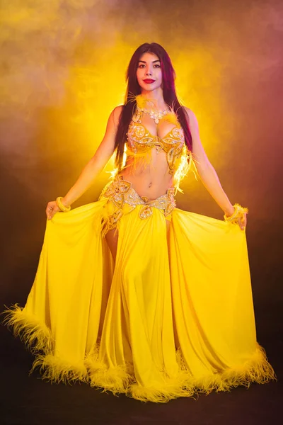Tempting sexy traditional oriental belly dancer girl dancing on yellow neon smoke background. Woman in exotic costume with feathers sexually moves her semi-nude body — Stock Photo, Image