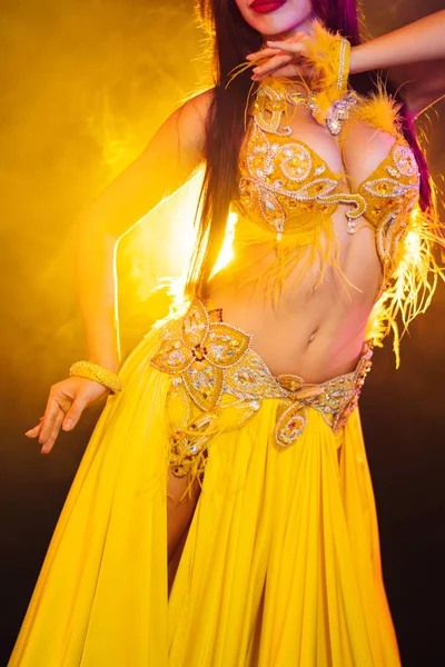 Närbild av traditionella orientaliska magdansös flicka dansa på gul neon rök bakgrund. Oigenkännlig kvinna i exotiska kostym med fjädrar flyttar sexuellt höfterna. — Stockfoto