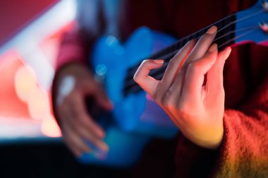 Eller genç güzel kız geceleri eğlence parkı içinde parlak neon merdivenlerde otururken mavi ukulele üzerinde oynama.