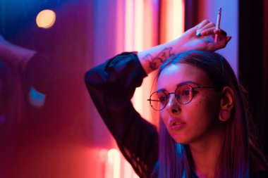 Bin yıllık gizemli güzel kızla gece parlak neon duvarının yakınında alışılmadık boyalı saç modeli. Saç, altın pullu çiller olarak mavi, piercing burun. Gizemli hipster genç gözlük