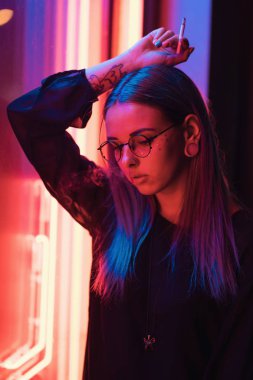 Bin yıllık gizemli güzel kızla gece parlak neon duvarının yakınında alışılmadık boyalı saç modeli. Saç, altın pullu çiller olarak mavi, piercing burun. Gizemli hipster genç gözlük