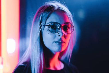 Bin yıllık gizemli güzel kızla gece parlak neon duvarının yakınında alışılmadık boyalı saç modeli. Saç, altın pullu çiller olarak mavi, piercing burun. Gizemli hipster genç gözlük