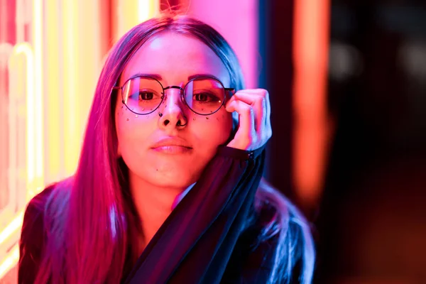 Menina bonita enigmática milenar com penteado tingido incomum perto da parede de néon brilhante à noite. Cabelo azul, lantejoulas douradas como sardas, piercing nasal. Misterioso hipster adolescente em óculos — Fotografia de Stock