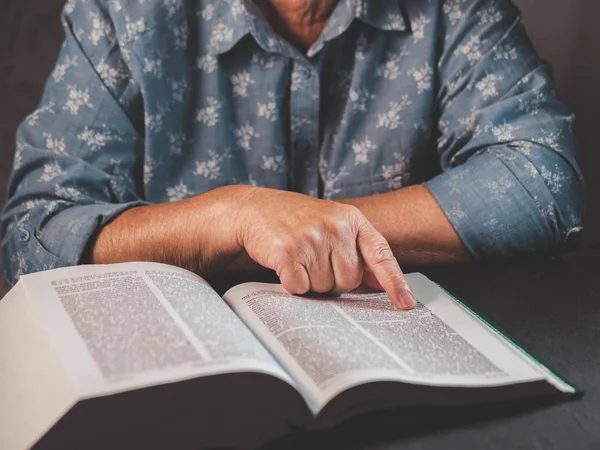 Old woman reading thick book at home. Grandmother with Bible. Concentrated elderly pensioner with wrinkles on hands attentively follows finger on paper page in library