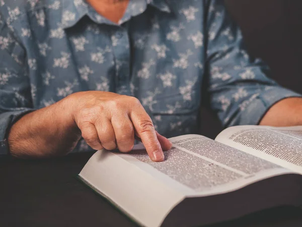 Una anciana leyendo un libro grueso en casa. Abuela con Biblia. Pensionista de edad avanzada concentrado con arrugas en las manos sigue con atención dedo sobre papel página en la biblioteca — Foto de Stock