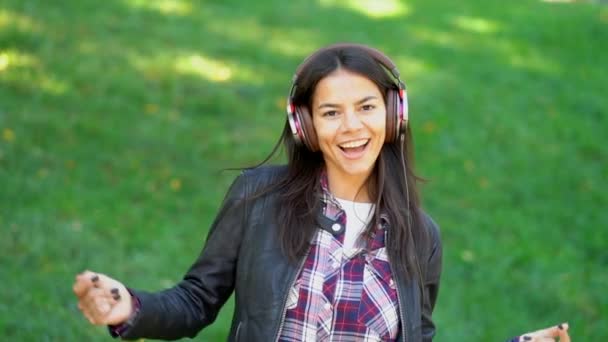 Mujer joven de raza mixta feliz escuchando música en los auriculares. Chica hipster hispana bailando al ritmo y cantando junto a la melodía en el parque — Vídeo de stock