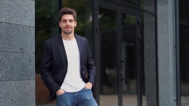 Retrato de un joven empresario exitoso y confiado en la ciudad en el fondo del edificio de oficinas. Hombre en traje de negocios mirando a la cámara y sonriendo. Retrato del chico guapo . — Vídeo de stock