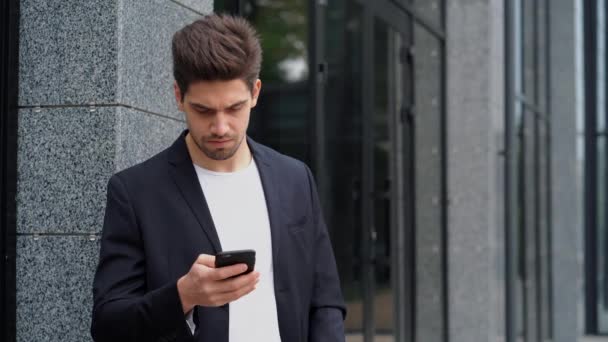 背景のオフィスビルで、スマート フォンの業務アプリを使用してアメリカのハンサムな男性。モバイル デバイス上の通信のスーツの青年実業家。4 k — ストック動画