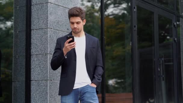 Americká pohledný muž používající obchodní aplikace na smartphone na kancelářskou budovu pozadí. Mladý podnikatel v obleku komunikaci na mobilním zařízení s sebejistý úsměv. 4k — Stock video
