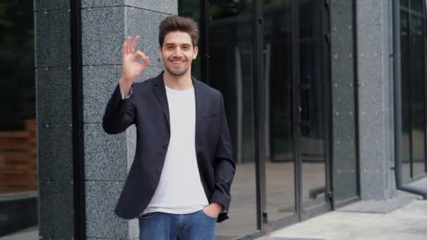 Successful happy man looking to camera and showing OK sign on office building background. Professional male manager wearing corporate suit jacket. Student guy outdoors. — Stock Video