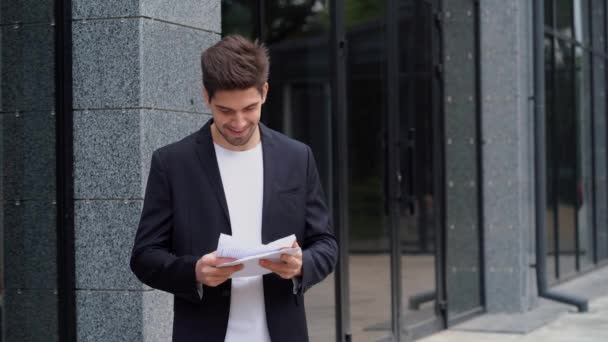 Mladý podnikatel hází papíry dokumenty do vzduchu a slaví úspěch na moderní kancelářská budova pozadí. Svoboda, úspěšné dokončení projektu koncept. — Stock video