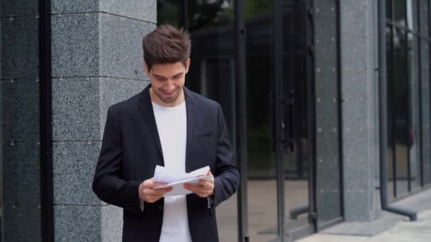 Joven hombre de negocios lanzando documentos de papeles en el aire y celebra el éxito en el fondo moderno edificio de oficinas. Libertad, finalización exitosa del concepto de proyecto . — Vídeo de stock