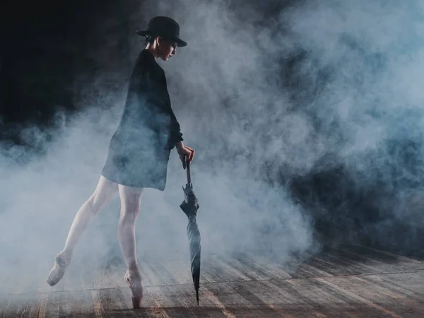 Bailarina em jaqueta longa preta e bodysuit sexy dançando no palco com luz mágica e fumaça. Jovem dançarina atraente em sapatos de ballet pointe com guarda-chuva — Fotografia de Stock
